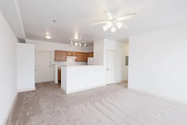 Living area facing the kitchen with white walls, neutral flooring, and a hallway on the right