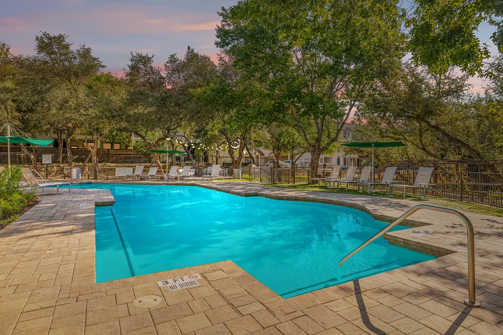 Community pool with lounge chairs surrounding, trees, and a black fence at dusk