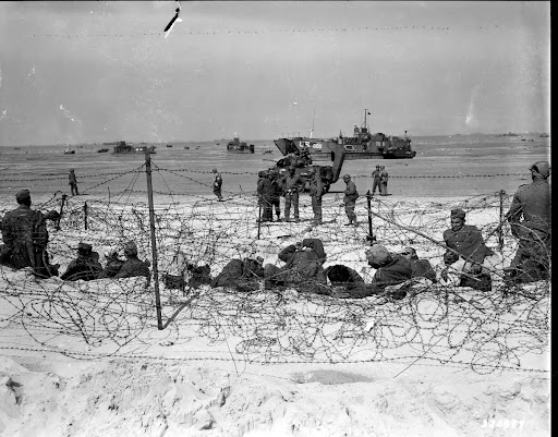 German prisoners rest in a barb-wired enclosure on Utah Beach after being interrogated by American soldiers. June 6, 1944.