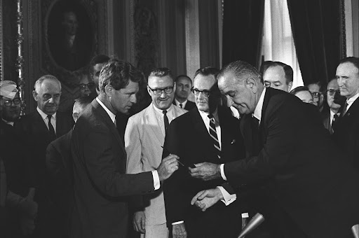 Pres. Lyndon B. Johnson handing a signing pen to Sen. Robert F. Kennedy as others look on.