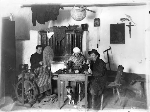 Pogled v "hišo". V ozadju peč. Moški kadi pipo. Ženska prede na kolovrat.