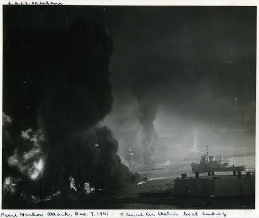 Streaming from shattered fuel tanks, oil turn parts of Pearl Harbor into a sea of flame, following the Japanese attacks. This picture was taken from near the Naval Air Station boat landing. Barely visible through the smoke are a damaged US Battleship and the capsized USS Oklahoma.