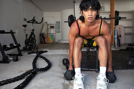 Bretman Rock sits at an exercise bench in a garage, holding weights in each hand. His chest is exposed, his yellow and black striped lycra shorts are visible. 