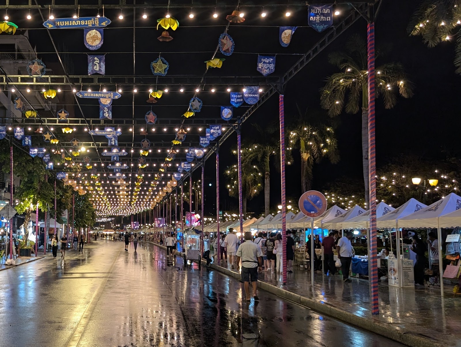 A street lit from above with string lights, with canopy tents
                 on along the right side shielding the street vendors from a
                 light drizzle. The street is empty except for folks walking
                 along the tents, and the light from above is reflected of the
                 wet pavement