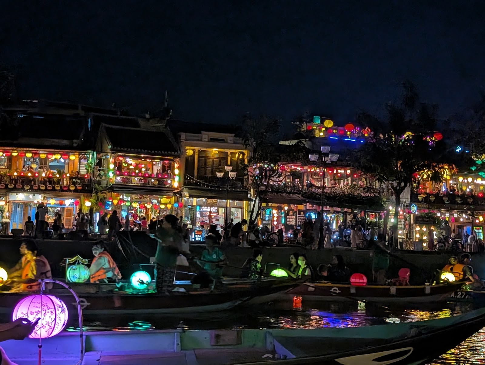 Photo with small canoe-style boats floating in a river in the foreground,
                 each with a different colored paper lantern. In the background, the river
                 bank is lined with people in front of a mostly yellow row of buildings,
                 with many paper lanterns hanging all over them.