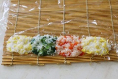 Trải một tấm tre dùng để cuốn cơm, trải thêm tấm màng bọc thực phẩm, múc mỗi loại cơm 1 chút đặt liền cạnh nhau cho lên theo thứ tự cơm trứng, cơm rau, cơm ớt rồi đến cơm trứng. Tiếp đó cuộn thât chặt cơm lại, vì có màng bọc sẽ giữ cho cơm vững.