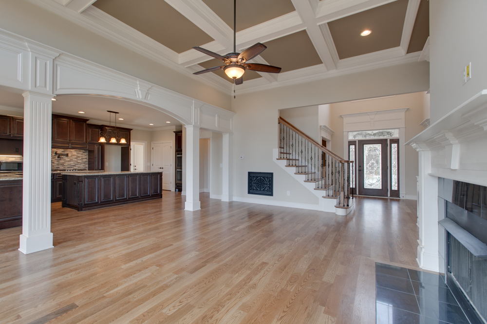An open-concept living space with hardwood floors, a coffered ceiling, a fireplace, a staircase, and an arched opening to a kitchen.