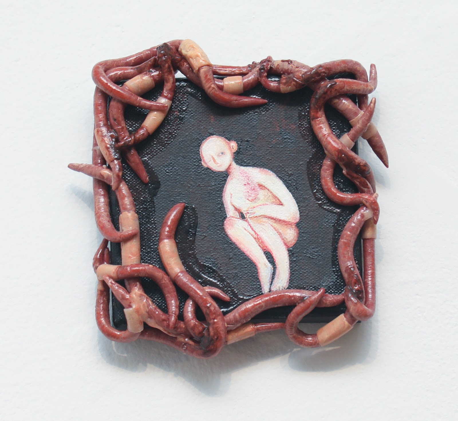 Small painted canvas with a sculpted frame. A pale, hairless figure crouches hunched over, hugging its knees, against a dark background. The frame is a tangle of glossy, worm-like tendrils sculpted from polymer clay in reddish-brown and flesh tones, with one tendril reaching into the painting toward the figure.