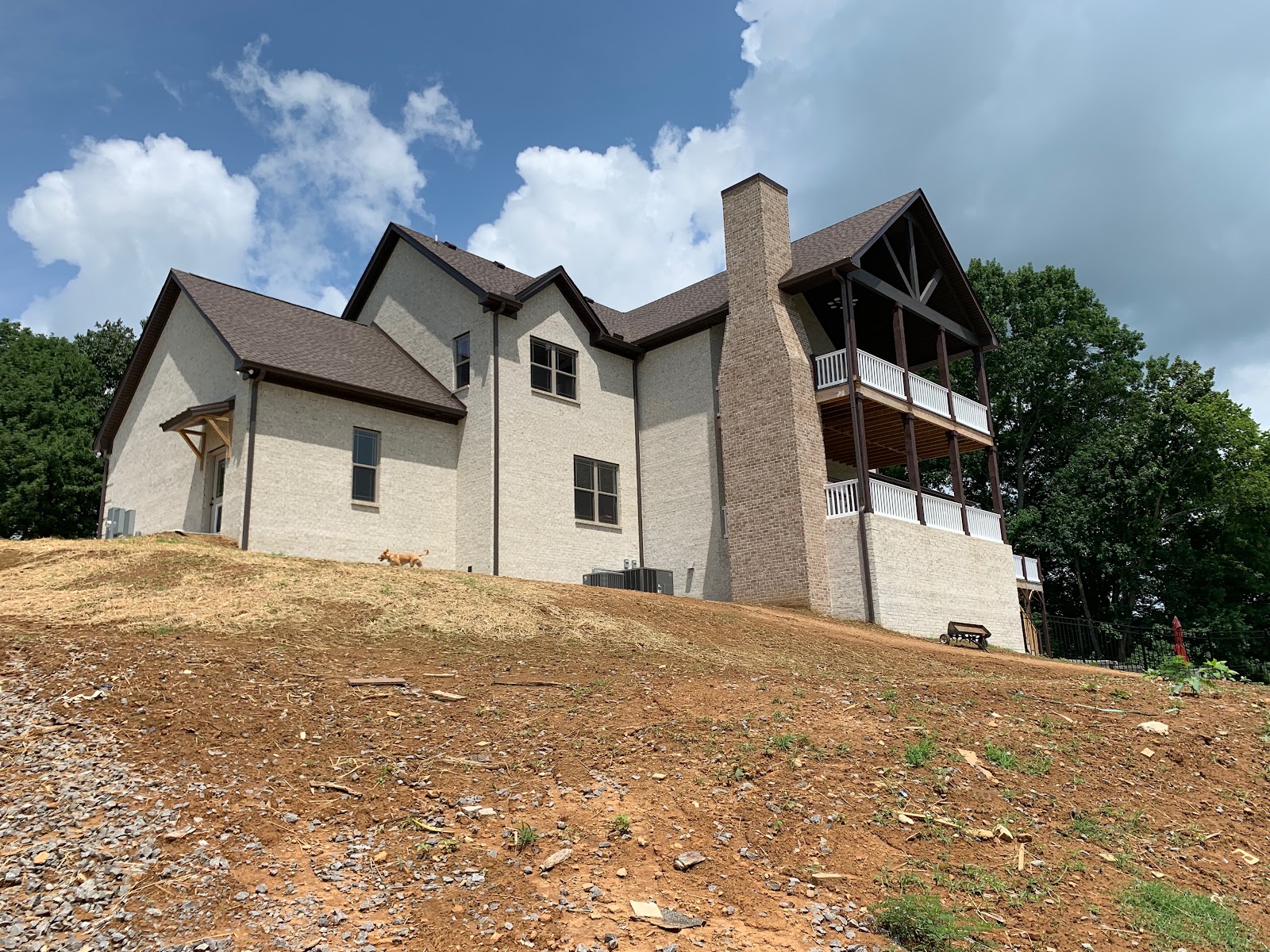 Side exterior view of a large light brick residence featuring a tall chimney and multi-level covered porches on a sloped dirt site.