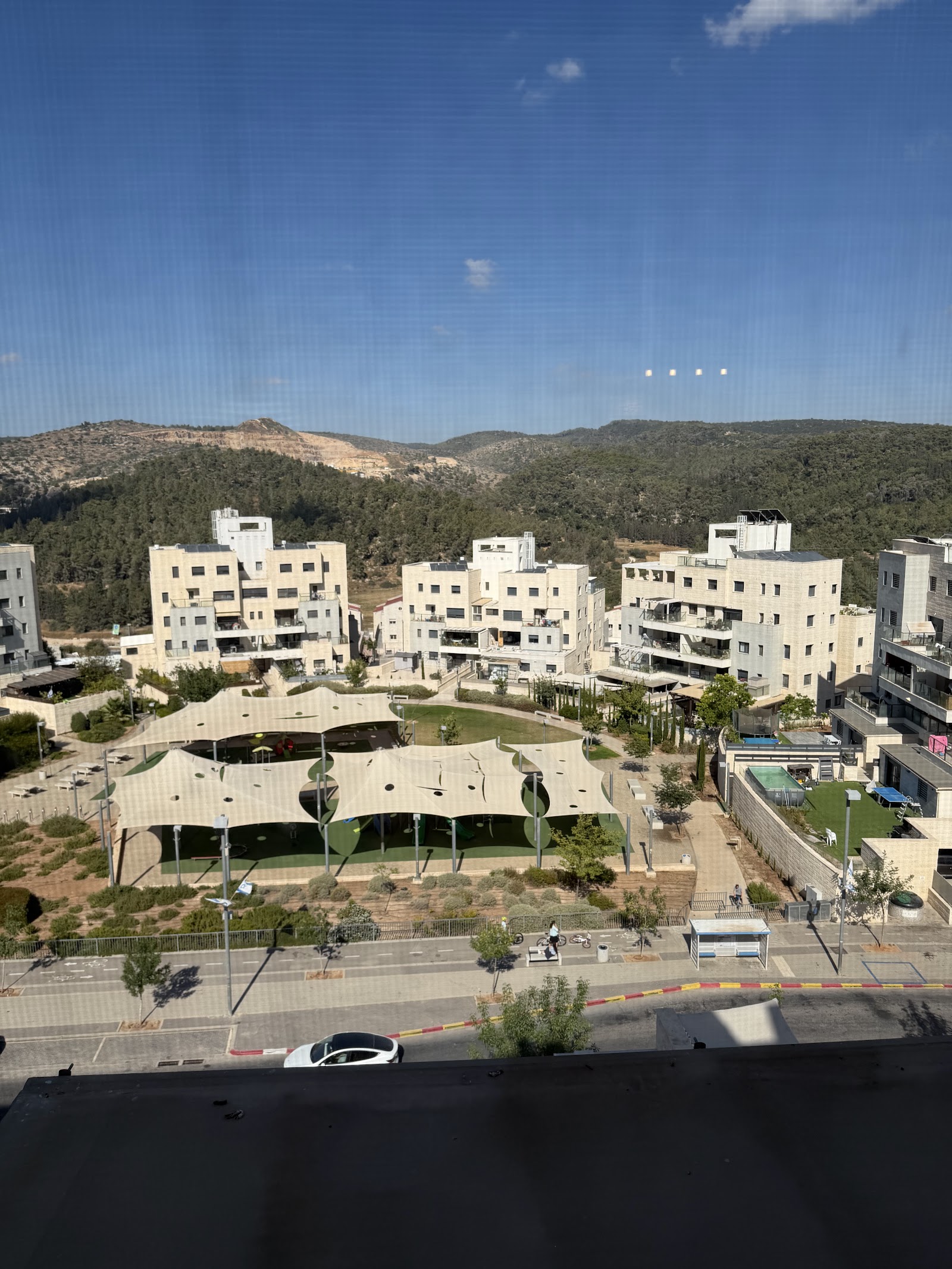 נוף שכונה בבית שמש מהחלון — בניינים בהירים, פארק וגבעות מיוערות