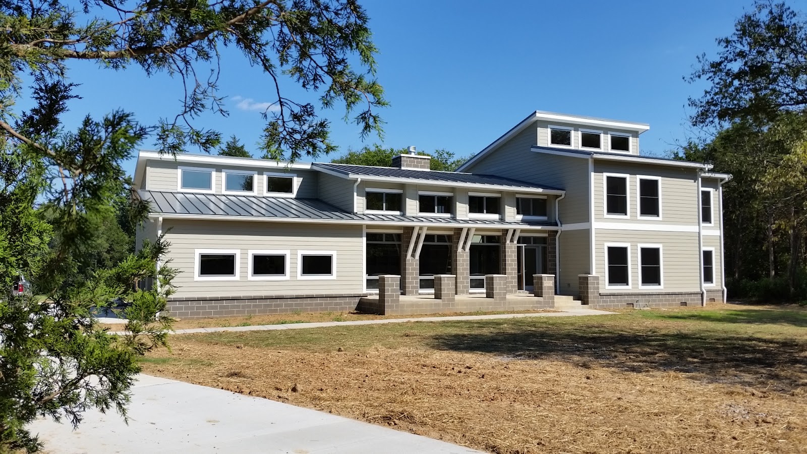 Exterior of a custom home with tan siding and a metal roof, showing masonry block pillars and a dirt yard, framed by tree branches in the foreground.