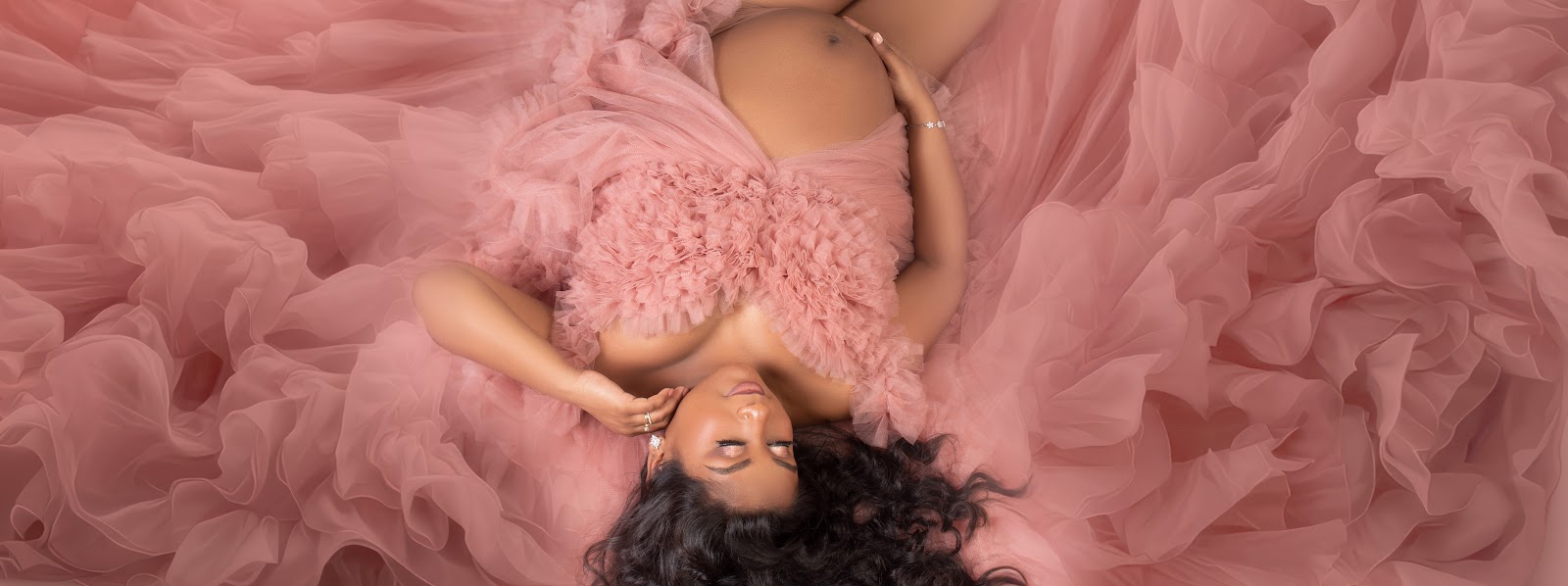 Studio maternity portrait from above: expectant mother lying on flowing blush-pink tulle, hand on belly, serene expression