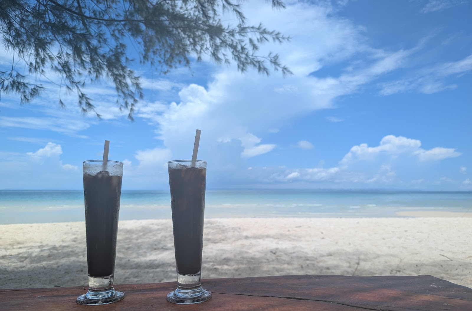 Two iced coffees on a wooden table in front of an empty white
                 sand beach, with turqoise water and a blue sky above