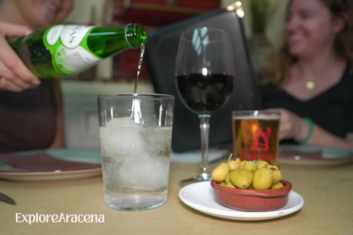 Sparkling water being poured beside red wine, beer and green olives, with two diners smiling in the softly blurred background.