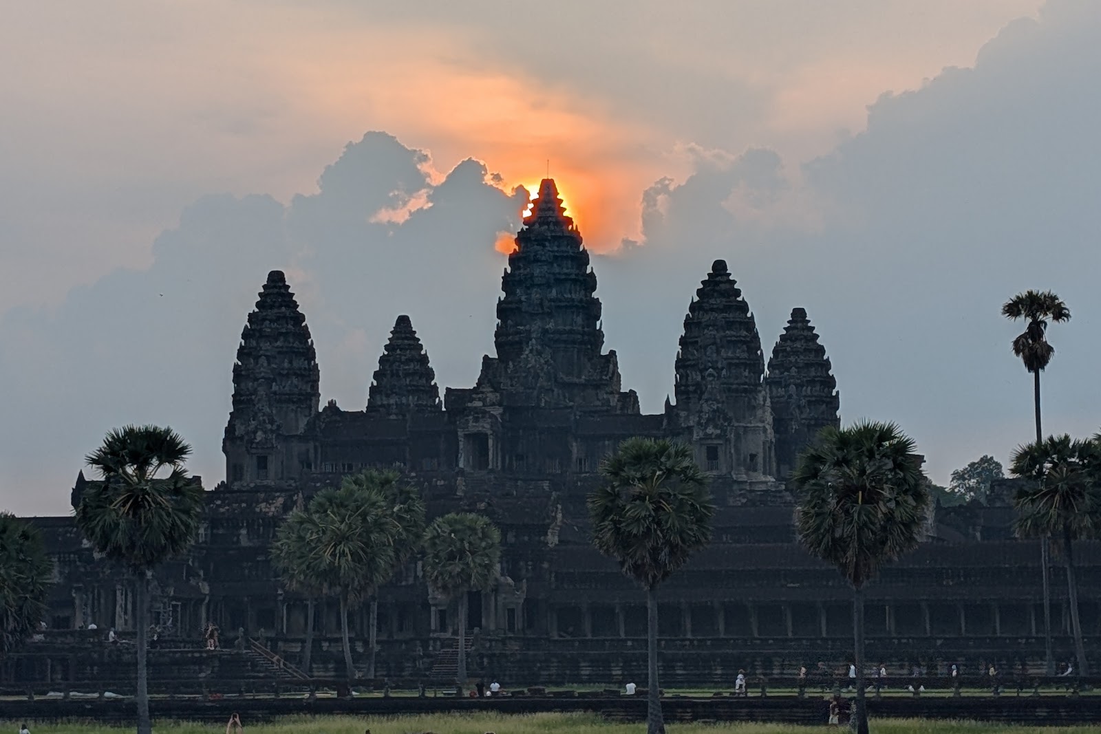 A slightly askew view of Angkor Wat with all five towers
                 visible, and the rising sun barely peaking over the top point.
                 Some palm trees are seen in the foreground.