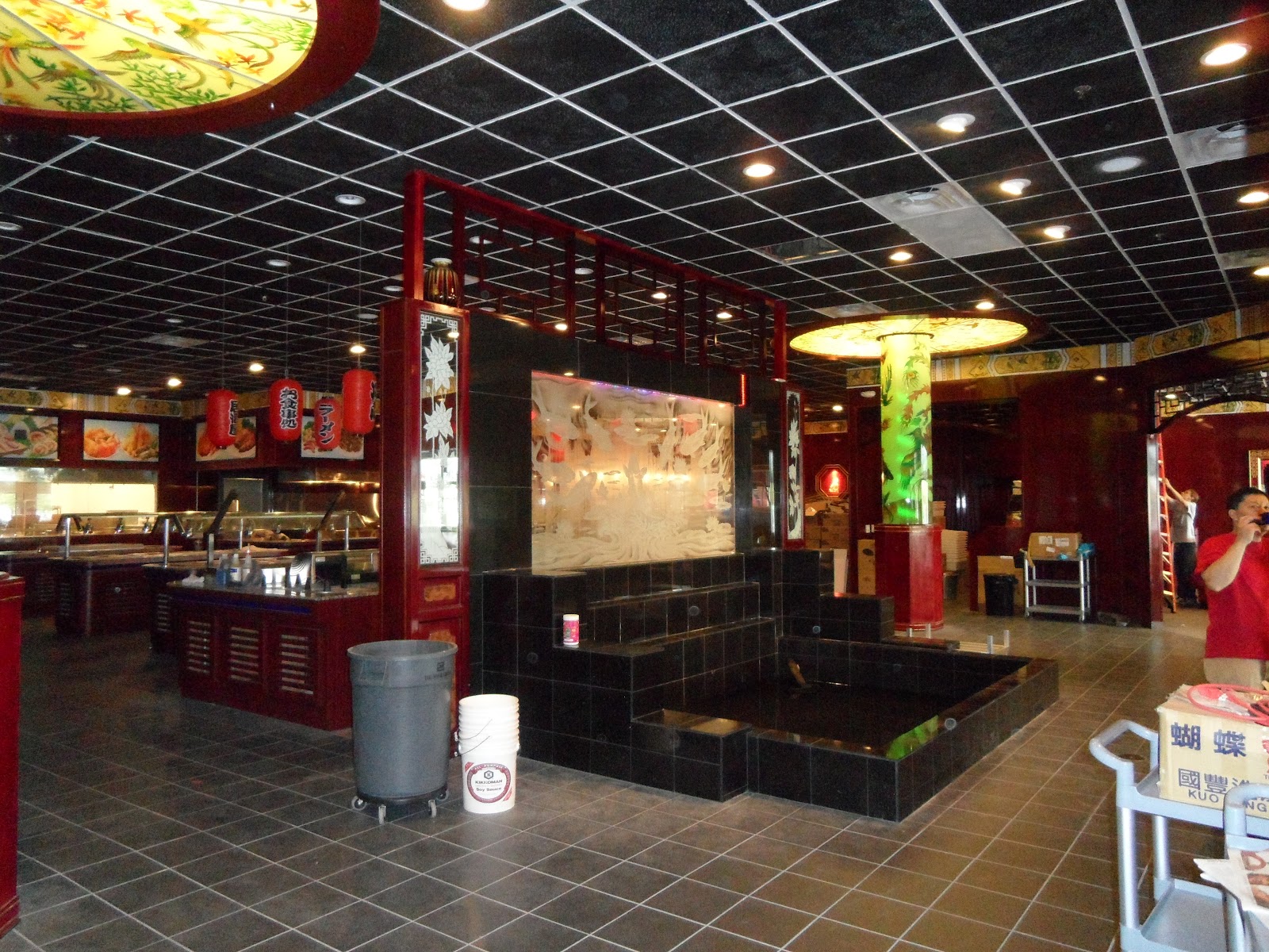Interior view showing a black tiled water feature, decorative columns with light fixtures, red lanterns, and buffet stations under a black grid ceiling.