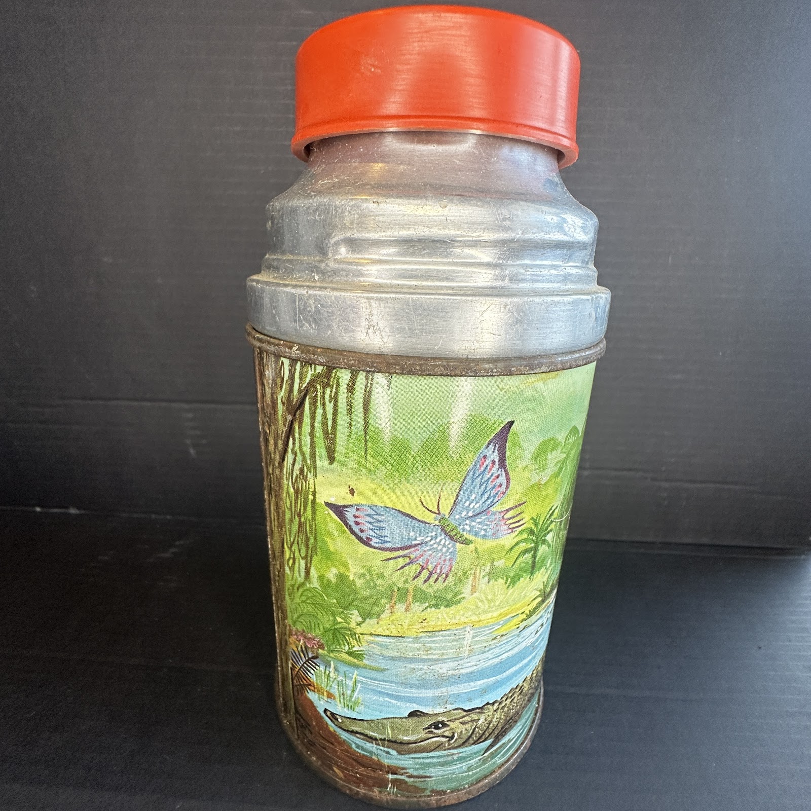 1977 Aladdin Industries metal thermos for Walt Disney's The Rescuers, showing lithographed swamp scene with Evinrude the dragonfly and crocodiles Brutus and Nero, red plastic cap, silver aluminum shoulder with rust on rims