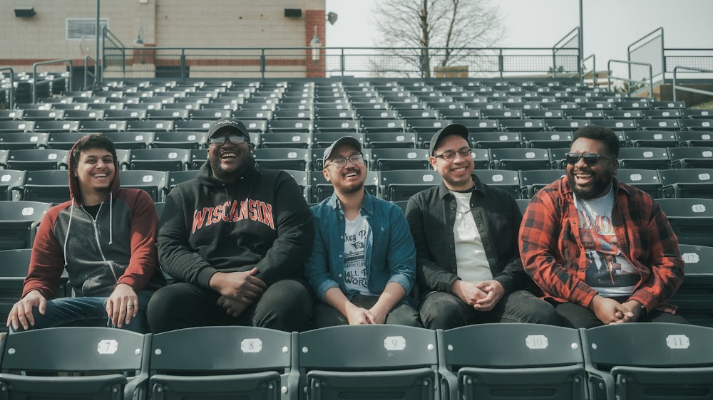 The five members of Action Adventure sit in a row in an outdoor sports arena