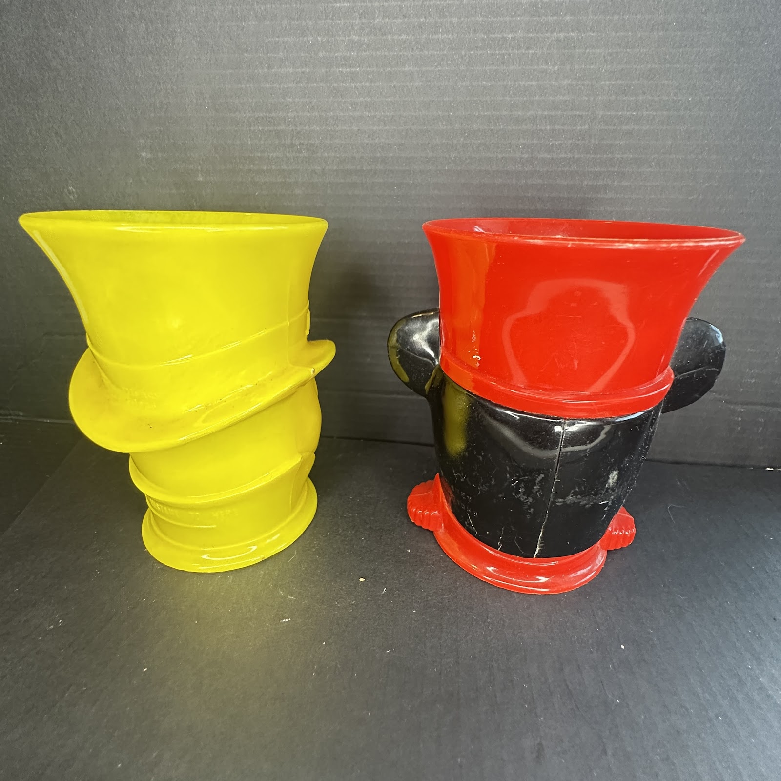 Set of two molded plastic Disney character souvenir cups: a solid yellow Donald Duck top-hat cup and a red and black Mickey Mouse ear cup, each approximately 6-7 inches tall, showing light age wear