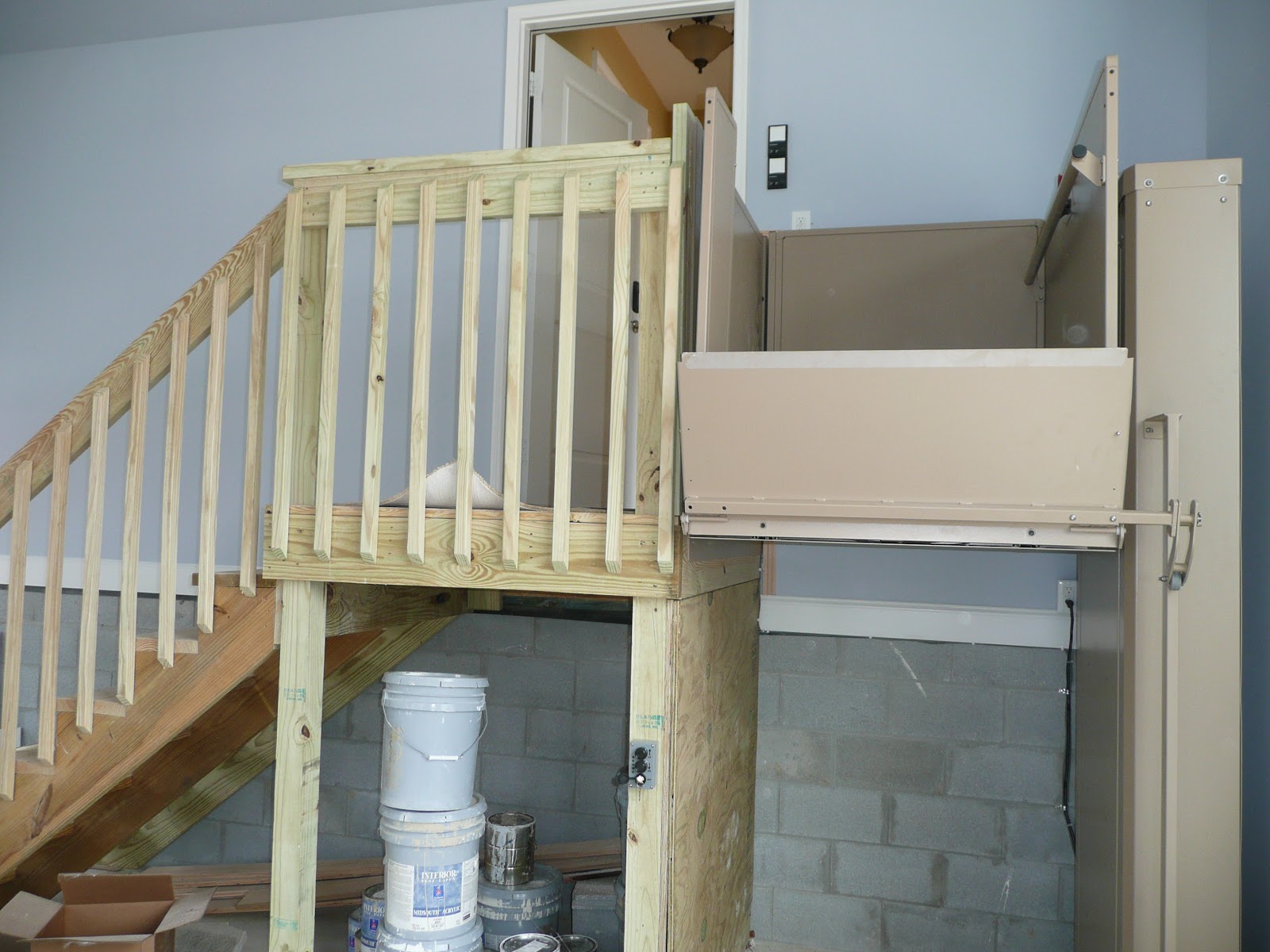 A beige platform lift in a raised position next to a wooden landing, with a wall-mounted central vacuum unit nearby.