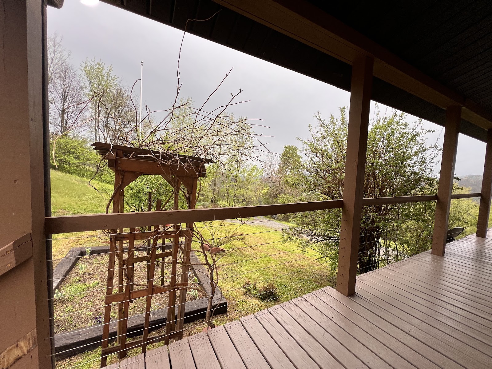 View from a wooden deck overlooking a grassy yard with a wooden trellis and surrounding trees under an overcast sky.