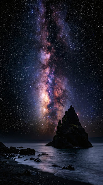 Vibrant glowing Milky Way galactic core arching over a dark monolithic coastal sea stack in pitch-black night.