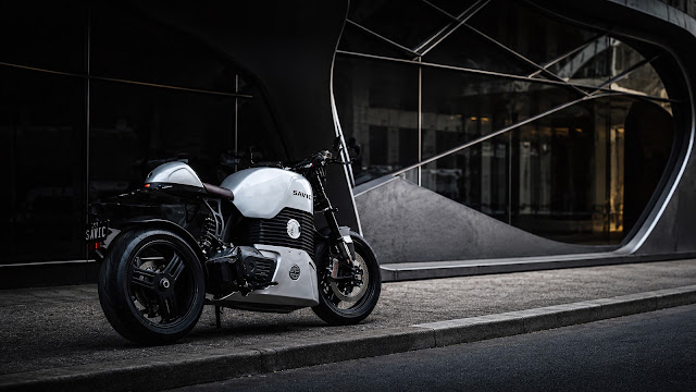 White Savic electric motorcycle parked on a concrete curb in front of an architectural modern building with large curved glass.