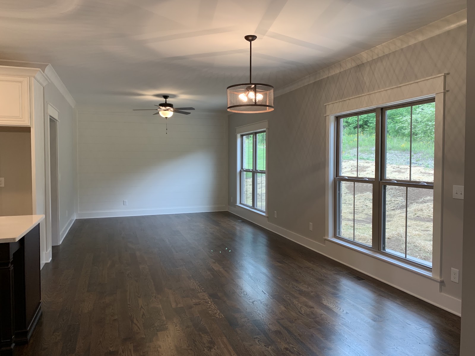 An empty room with dark wood floors, light gray walls, two large windows, a ceiling fan, and a drum-style chandelier.