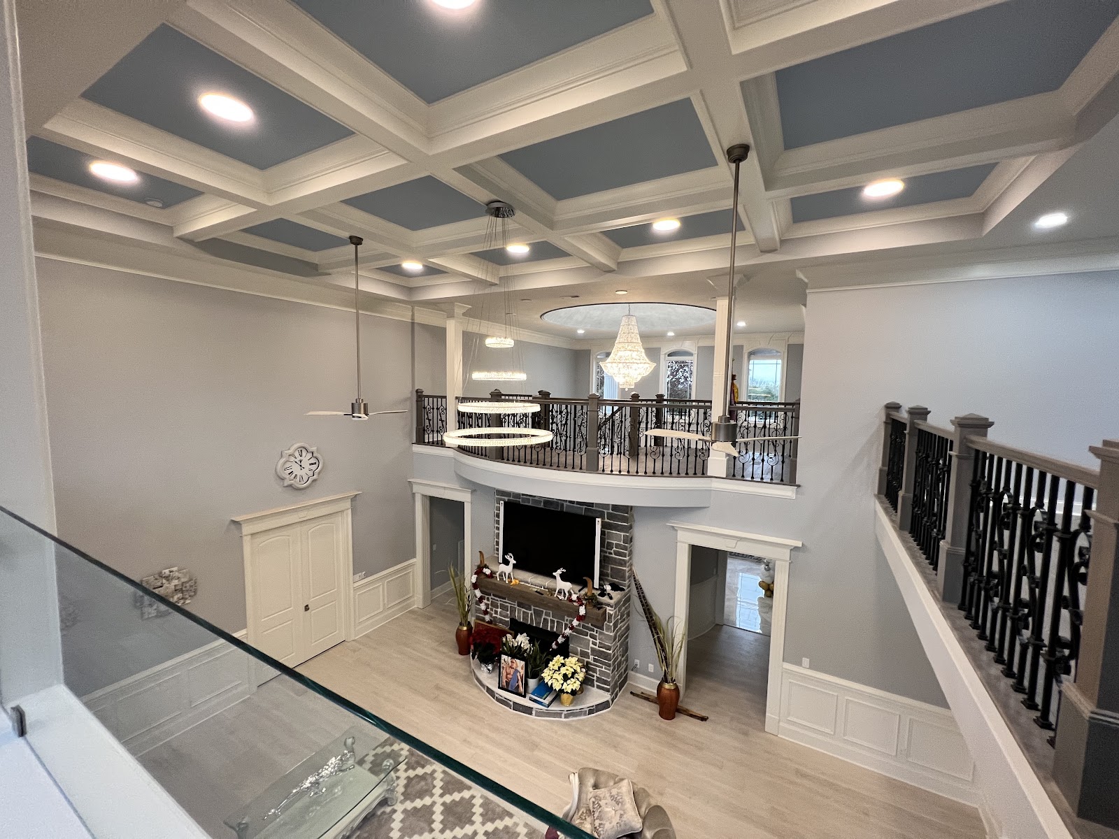 High-angle view of a two-story living area with a coffered ceiling, stone fireplace, and an upper-level balcony with dark railings.
