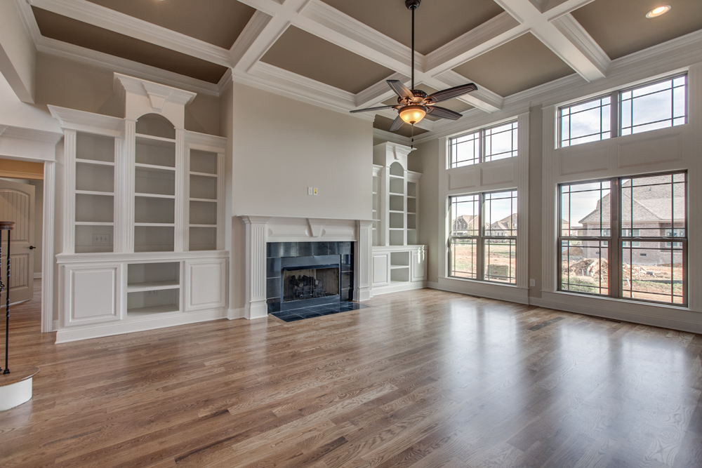 Living area in a Marymont residential construction project featuring a coffered ceiling, built-in white shelving, a fireplace, and large windows.