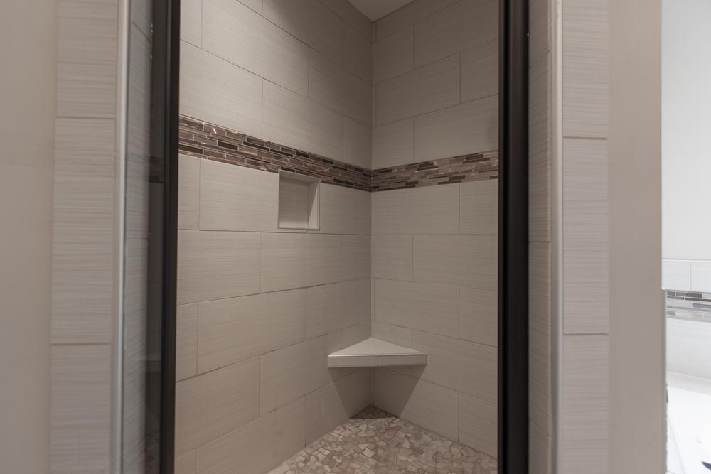 Interior view of a tiled shower stall with a corner bench, recessed wall niche, and a decorative mosaic tile band. The floor features small, irregular stones.