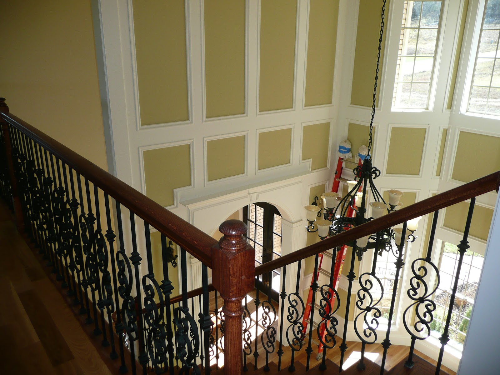 Interior view from a balcony showing a wooden handrail with wrought iron balusters, decorative wall paneling, a chandelier, and an orange ladder.