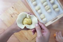 Đặt viên nhân bánh pía vào giữa miếng bột vỏ, cho trứng muối vào rồi gói lại cho kín nhân, dùng tay nắn nhẹ để được khối cầu đều. Bạn cần gói cho lớp vỏ bánh pía Sóc Trăng bám sát viên nhân bánh, dính mép bột chặt tay để nhân không bị hở ra ngoài khi nướng. Phần bột thừa ra bạn dùng tay túm chặt lại giống như túi gói quà. Quay phần túm bột xuống dưới làm đế bánh pía Sóc Trăng. Đặt bánh đã nặn xong lên khay nướng có lót giấy nến chống dính. Dùng tay đè cho bánh pía Sóc Trăng hơi dẹt xuống, bề mặt bánh bằng phẳng.