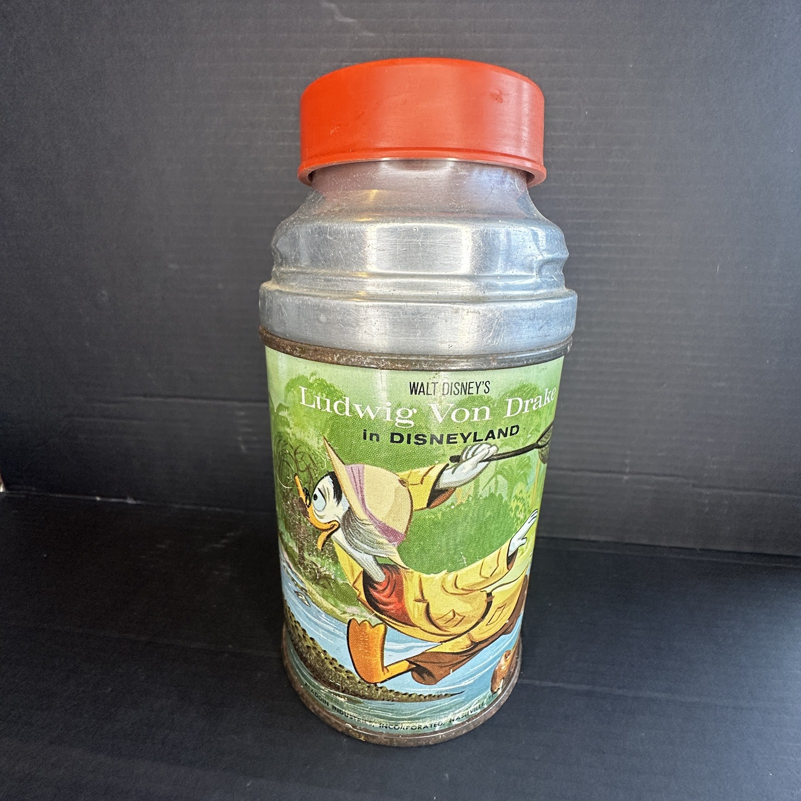 Vintage Aladdin Industries lithographed tin thermos circa 1961–1963 showing Ludwig Von Drake in yellow rain slicker and pith helmet carrying a butterfly net, with red plastic screw-on cap, wear and oxidation consistent with age