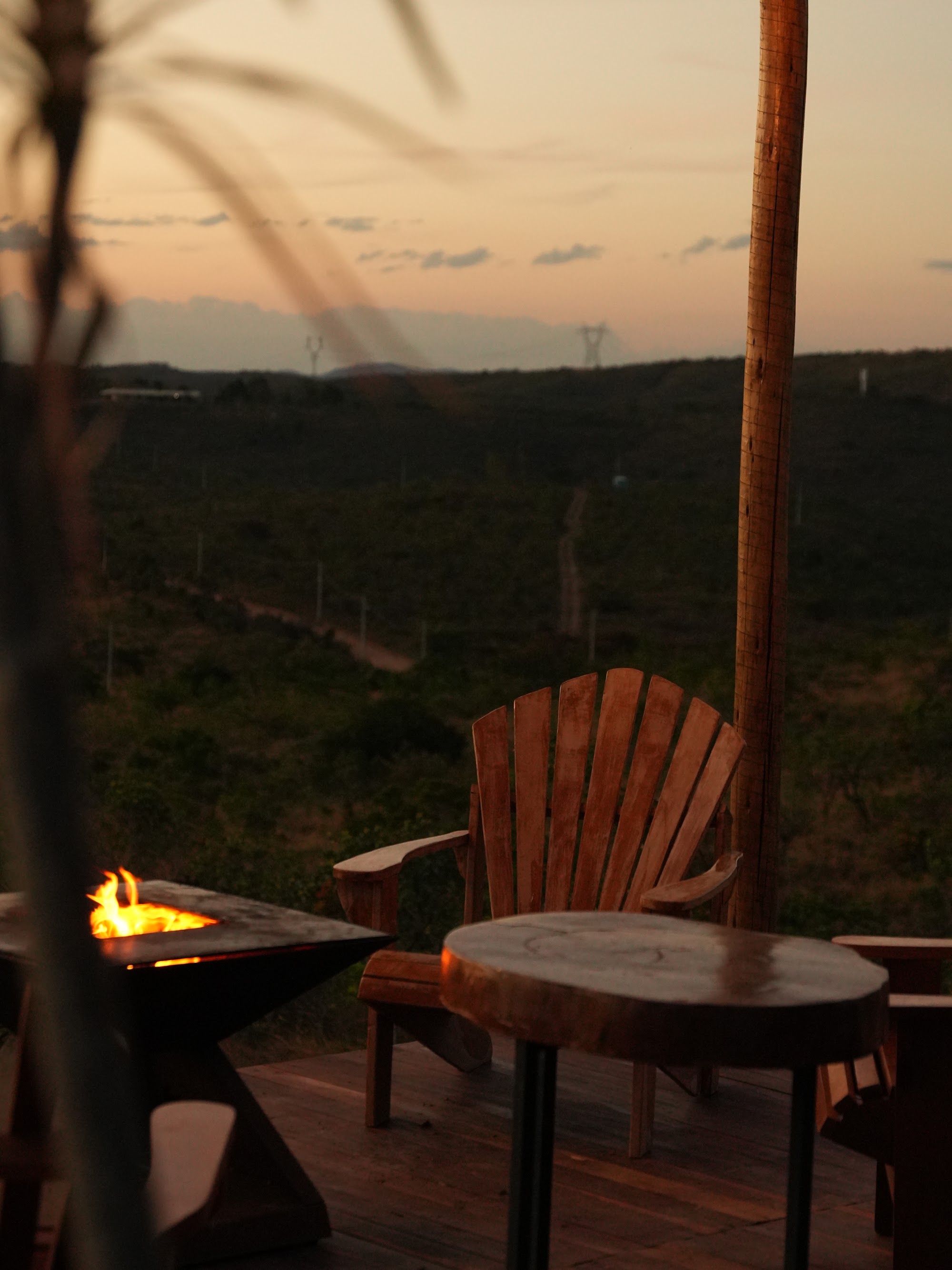 Cadeira de madeira e pit fire aceso no deck de um chalé ao entardecer, com vista aberta para o campo e a serra ao fundo