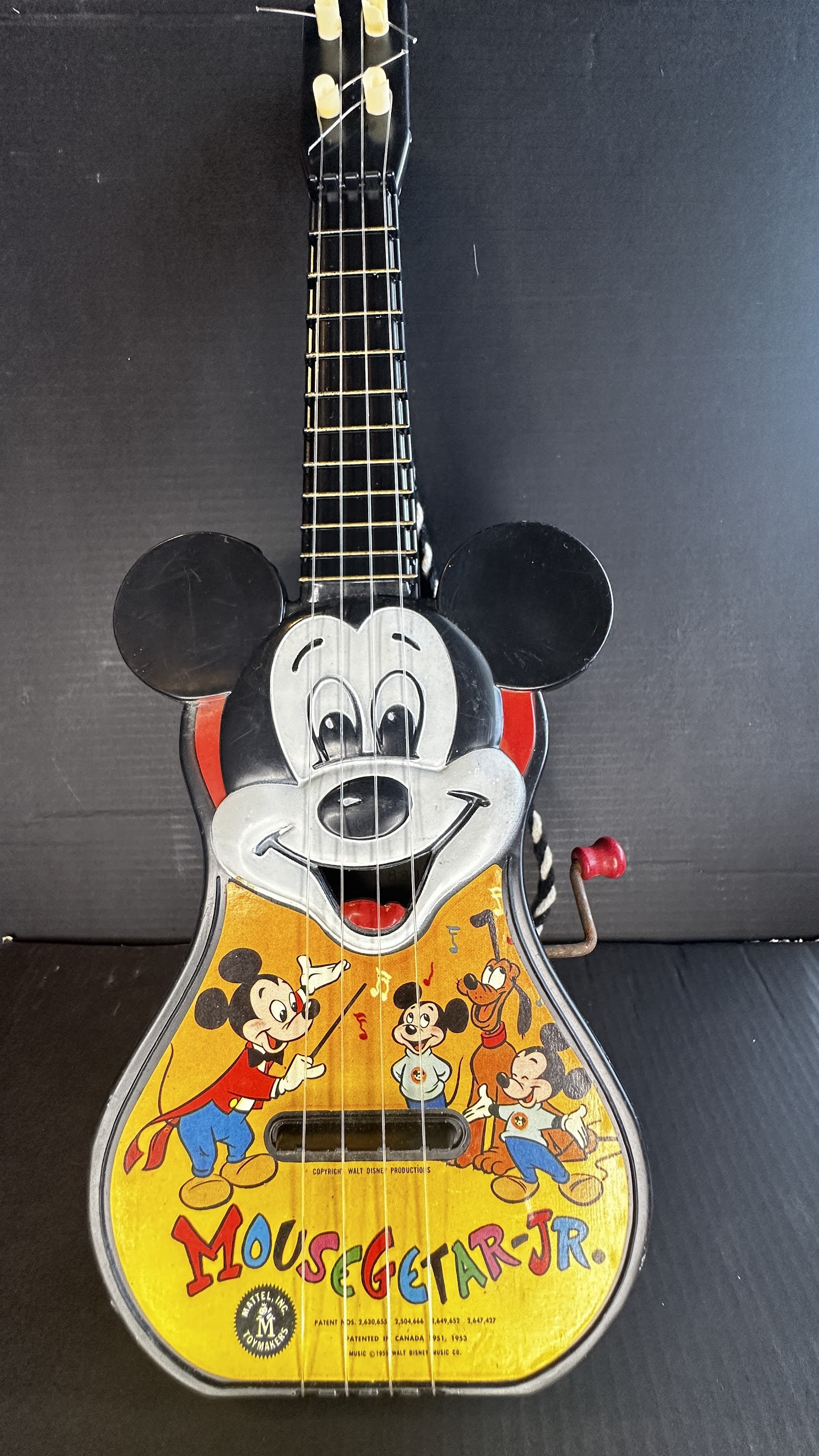 Vintage Mattel Mousegetar Jr. toy ukulele with black Mickey Mouse ear body, yellow soundboard showing Mickey, Pluto, and Morty and Ferdie Fieldmouse, and side hand-crank music box mechanism, circa 1955–1958