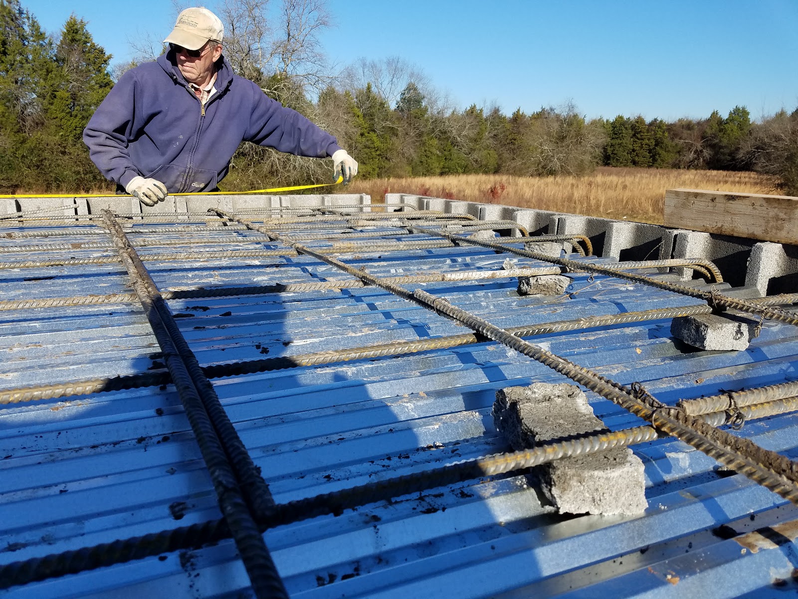 A worker uses a measuring tape on a rebar grid laid over a metal deck for a storm shelter roof.