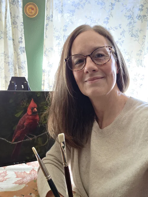 Kelly Allerd holding paint brushes in front of a picture of a red bird