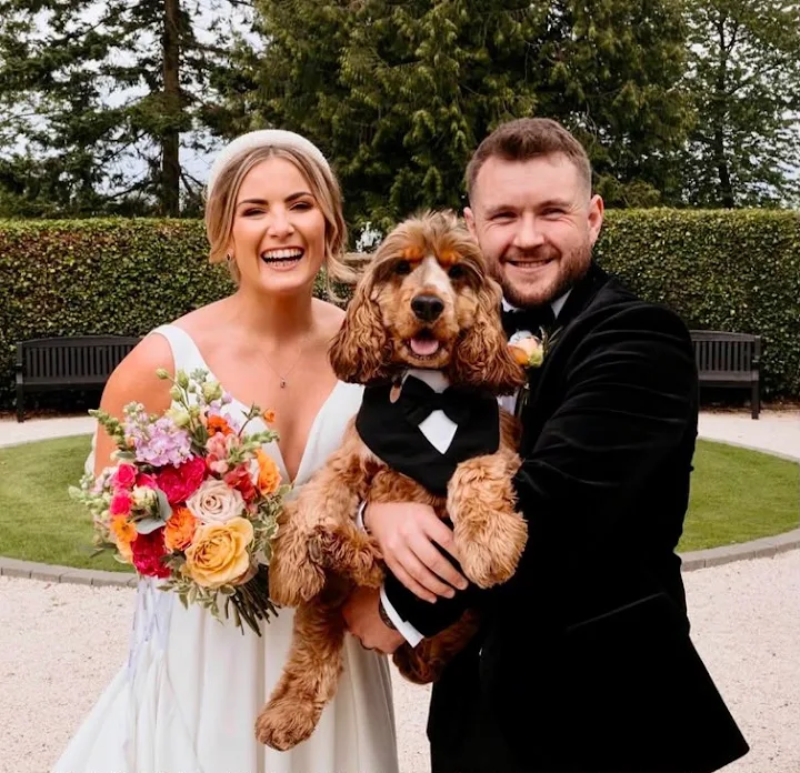 A bride and groom laughing with their cockapoo in a bow tie on their wedding day in Wiltshire