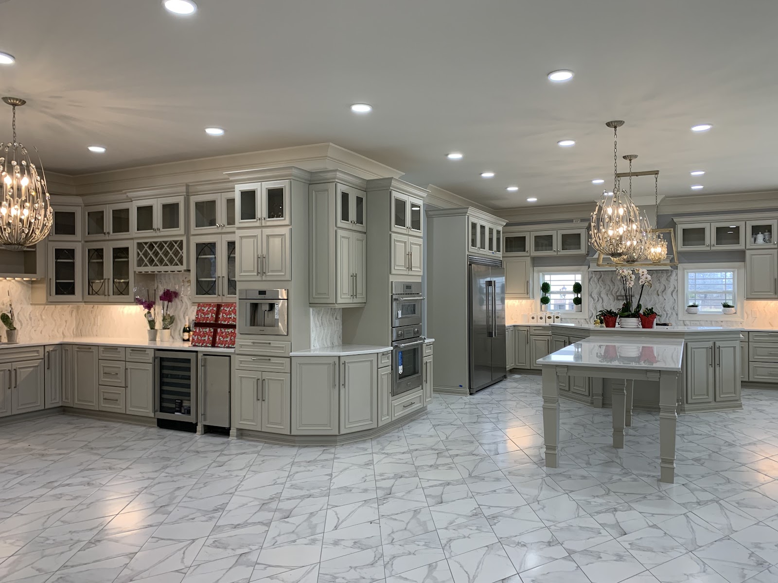 Spacious kitchen featuring light gray cabinetry, marble-patterned tile flooring, two large crystal chandeliers, and a central island with a white countertop.
