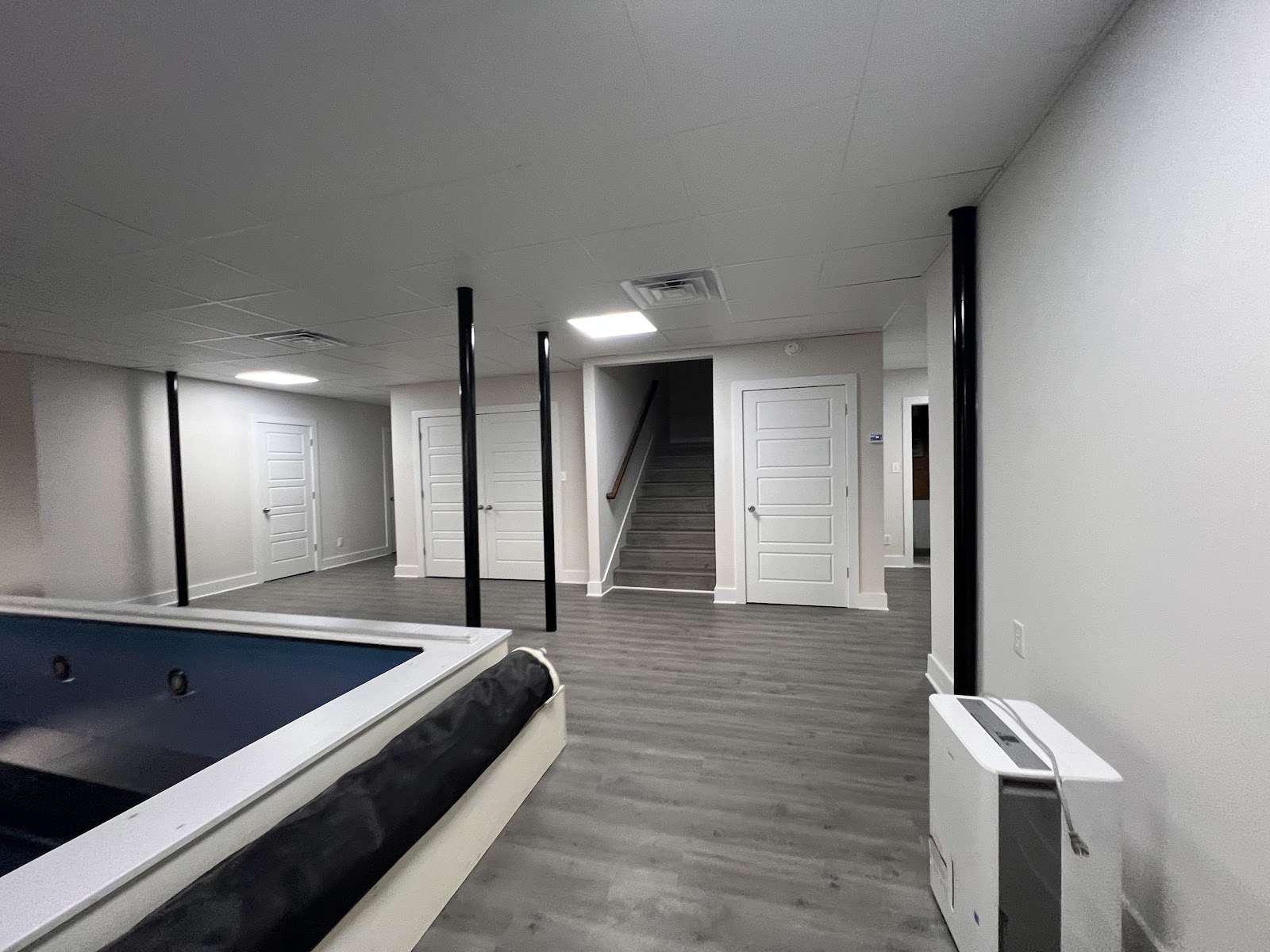 Indoor space with grey flooring, white walls, and black support columns. A blue pool with a black cover is in the foreground. White doors and a staircase are visible in the background.