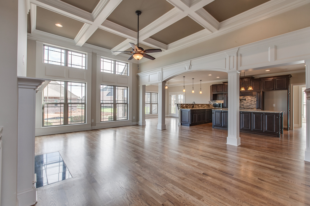 Interior living area with hardwood floors, a coffered ceiling, and large windows, looking through an arched opening into a kitchen with dark cabinetry.