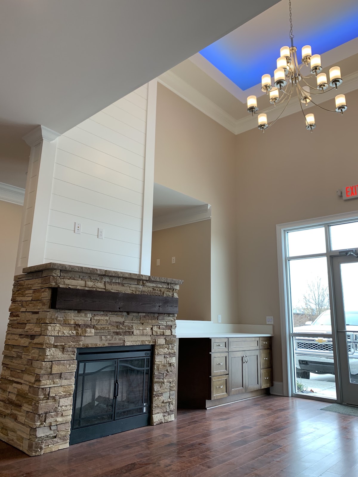 Interior room with a stone-faced fireplace, dark wood mantel, and built-in desk with brown cabinets. A chandelier hangs from a recessed ceiling with blue accent lighting.