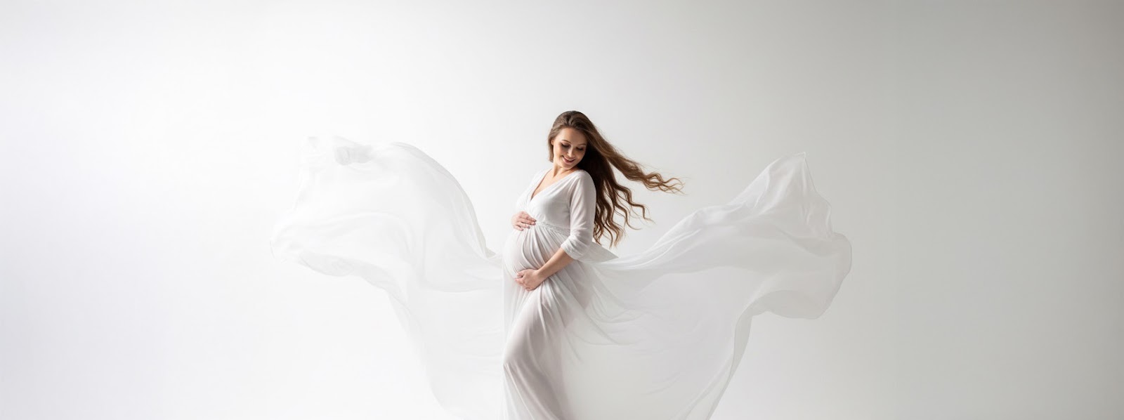 Studio maternity portrait: pregnant woman in a white gown with dramatic flowing chiffon creating wing-like shapes, side profile, hand on belly, white backdrop