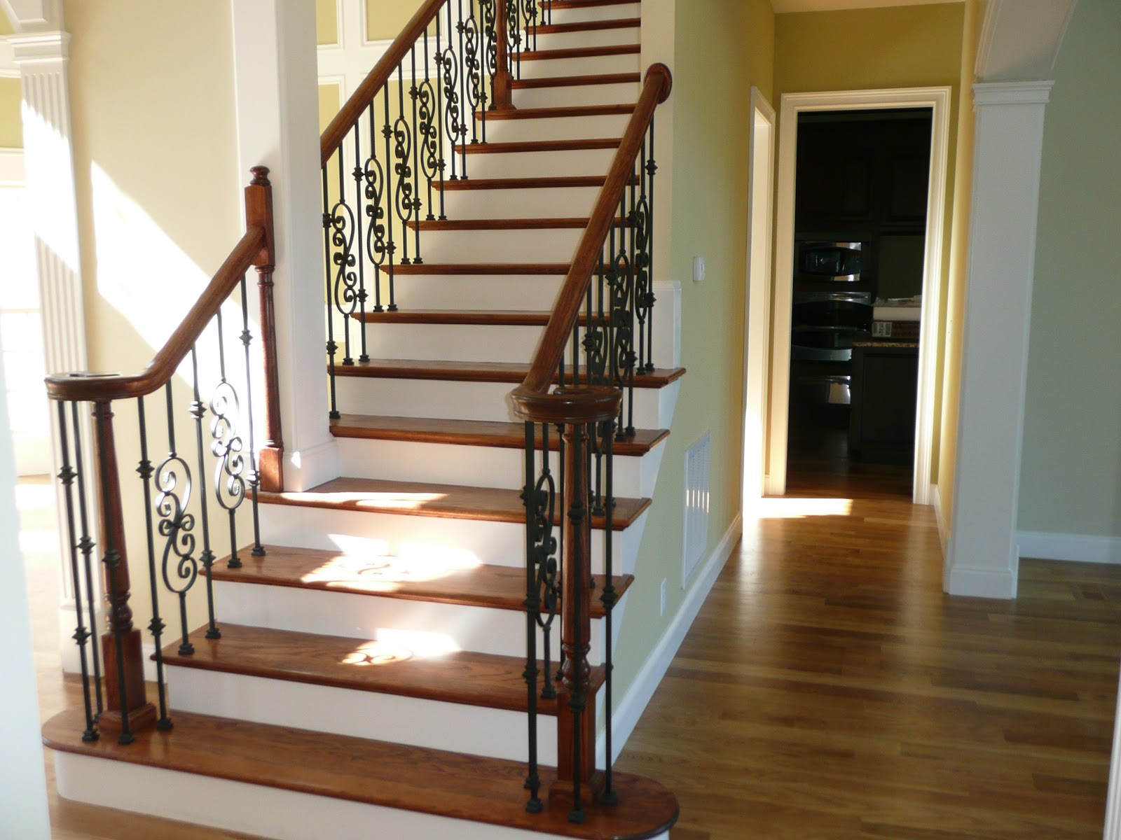 Interior view of a staircase with wooden treads and handrail, decorative black metal balusters, and hardwood flooring.