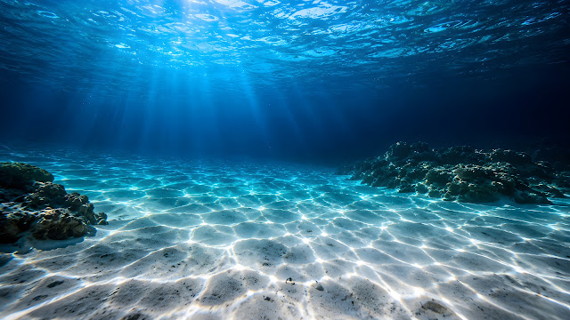 Underwater view of crystal clear blue ocean water with bright sunbeams dancing on white sandy seabed.