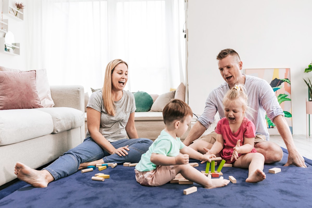 Family playing with toys on blue play mat in bright living room with couch