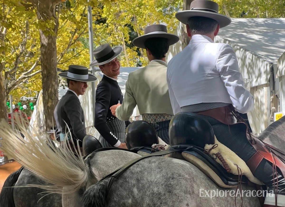 Four young men riding horses together, with one turning back and smiling during the Feria de Aracena.