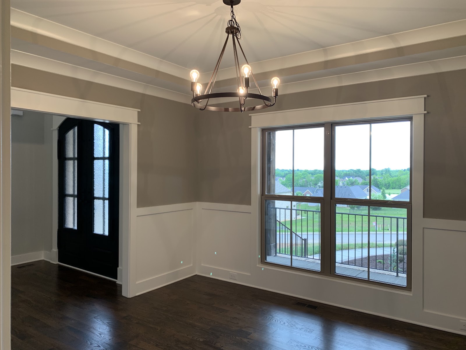 Room with dark wood floors, gray walls with white wainscoting, a tray ceiling with a black chandelier, and a large window overlooking a neighborhood.