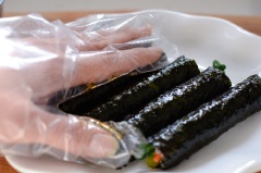 Rong biển tấm bự bạn cắt làm tư rồi trải ra mặt phẳng, cho một lớp cơm mỏng lên trên rồi đặt củ cải muối, cà rốt, cải bó xôi lên và cuộn chặt tay lại. Làm như vậy cho đến hết nguyên liệu. Làm nước sốt: cho nước tương, giấm gạo, đường, mù tạt vàng và chút nước vào chén khuấy đều. Vậy là chúng ta hoàn thành rồi đó.